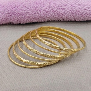 Peut inclure: Un ensemble de cinq joncs fins en or, chacun présentant un motif gravé délicat et répétitif de fleurs ou de feuilles sur la surface extérieure. Les bracelets sont circulaires, métalliques, avec une finition polie, empilés en se chevauchant.