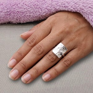 May include: A silver ring featuring a large, rectangular face with a hammered, textured surface. The metal has a polished, reflective finish that catches the light, creating a distinct pattern across the flat, slightly curved rectangular plate.