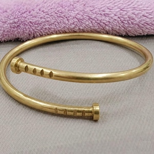 Könnte beinhalten: Ein goldfarbenes, offenes Armband mit Nagelkopfdessign. Das Armband hat ein gebogenes, zylindrisches Band mit einer Reihe kleiner, gleichmäßig verteilter Vertiefungen. Die Enden des Armbands haben eine abgerundete Nagelkopfform. Das Armband wird auf einer hellgrauen Oberfläche präsentiert.