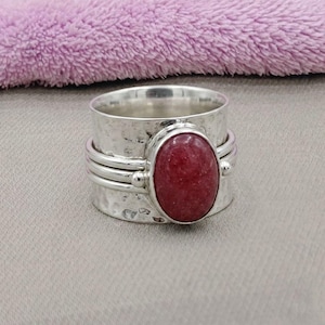 Könnte beinhalten: Ein breiter Silberring mit Drehfunktion, der eine gehämmerte Textur auf dem Band und einen ovalen roten Edelstein in der Mitte aufweist. Der Ring besteht aus einer breiten Basis mit zwei darüber liegenden, schmaleren Silberringen um den Stein.