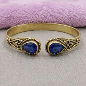 Può includere: Un bracciale aperto color oro con due pietre blu scuro a forma di goccia alle estremità. La fascia in metallo è decorata con intricate texture a corda e piccoli accenti circolari che circondano ciascuna delle due gemme blu.