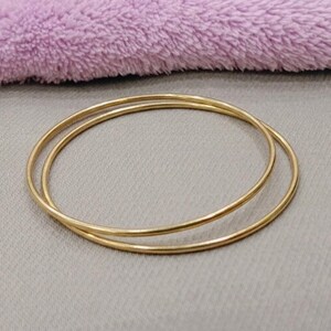 Può includere: Due braccialetti rigidi circolari e sottili in metallo color oro con una finitura liscia e lucida. I bracciali sono semplici, privi di decorazioni e presentano un design continuo e senza giunture. Questi accessori metallici sono leggeri e hanno una superficie riflettente.