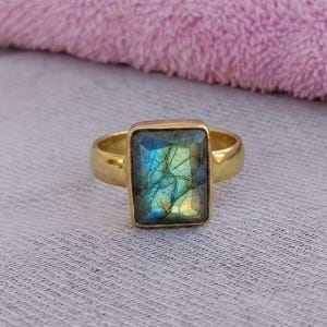May include: A gold-colored ring featuring a rectangular labradorite stone. The stone displays a mix of blue, green, and yellow hues. The ring is set on a light gray surface with a pink towel in the background.