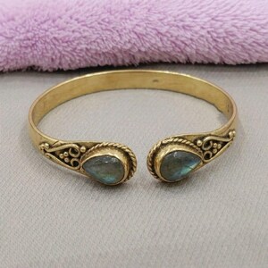 May include: A gold-toned open cuff bracelet featuring two teardrop-shaped labradorite stones at the ends. The metal band is decorated with intricate wire-twisted scrollwork and small bead accents surrounding each stone setting.