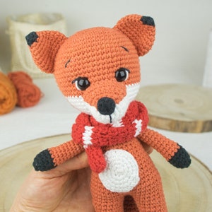 May include: A crocheted fox plush toy with a red and white striped scarf. The fox has black eyes and a black nose. The toy is sitting on a wooden surface.