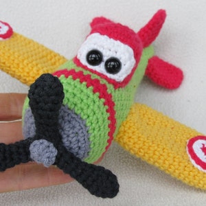 May include: A handmade crochet airplane toy in bright colors. The toy features a green body, red and white accents, yellow wings, and a black propeller. The airplane has large, black button eyes and a red and white hat.