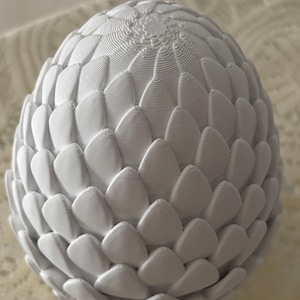 May include: A white, egg-shaped ornament with a textured surface. The surface is covered in overlapping, petal-like shapes, creating a layered effect. The top features a spiral design. The object is likely made of a solid material.