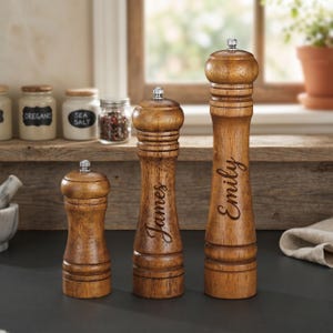 May include: Three wooden pepper grinders of varying sizes. The grinders are a warm brown color with a silver top. The names "James" and "Emily" are engraved on two of the grinders. The grinders are on a kitchen counter.