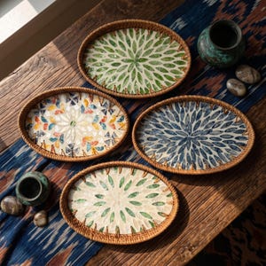 Puede incluir: Cuatro bandejas ovaladas con bordes de ratán tejido e incrustaciones de mosaico de conchas. Los diseños incluyen motivos de pétalos florales en verde, azul y multicolor dispuestos en patrones radiales sobre un fondo claro.