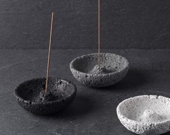 May include: Three circular incense holders with a rough, porous texture. One holder is black, one is dark gray, and one is light gray. Each bowl-shaped holder features a small central mound with a hole to hold a thin, brown incense stick upright.