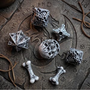 May include: A set of seven polyhedral dice with a weathered, bone-like texture and dark ink numbering. The collection includes various geometric shapes, including a spherical die and two bone-shaped pieces, all featuring a distressed white finish.