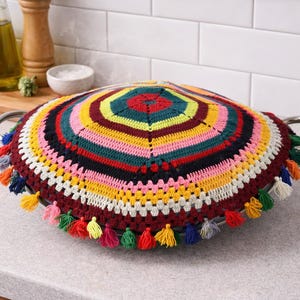 Puede incluir: Una cubierta de tapa de olla de ganchillo multicolor con un diseño circular. La cubierta presenta una gama de colores, incluyendo rojo, amarillo, verde y rosa, y está adornada con borlas decorativas. La tapa está colocada sobre una olla de metal.