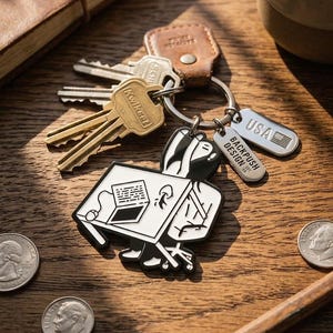 May include: A metal keychain featuring a black and white enamel charm of a person sitting at a desk with a laptop. The keychain includes a brown leather tab, several metal keys, and two rectangular tags stamped with USA and BACKPUSH DESIGN EST.
