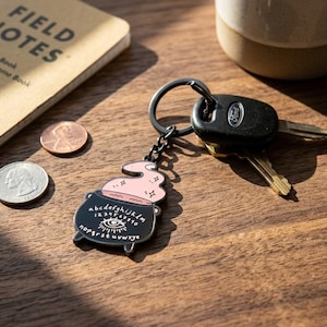 May include: A black metal keychain featuring a cauldron-shaped charm with a pink swirling liquid top. The cauldron is printed with the alphabet, numbers 0-9, and an eye symbol. It is attached to a black metal split ring holding a black car key.