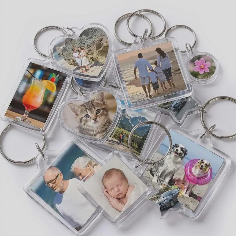 May include: A collection of clear acrylic keychains in various shapes including hearts, squares, and stars, each featuring a different printed photograph such as a beach scene, a kitten, a cocktail, a baby, and a dog with a pink floatie.