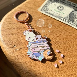 May include: A rose gold metal keychain featuring a white rabbit character leaning over a light purple cauldron filled with swirling liquid. The design includes a crescent moon, small stars, and the text Seven Art. A one-dollar bill is visible nearby.