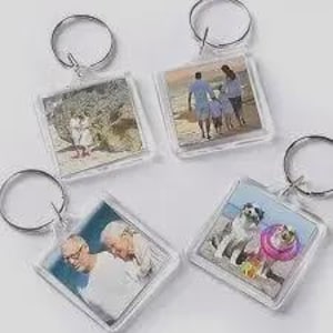 May include: Four square acrylic keychains, each featuring a different photograph insert. The images include a couple on a beach, a family walking on the sand, two older individuals, and two dogs, one wearing a pink collar, all attached to metal rings.