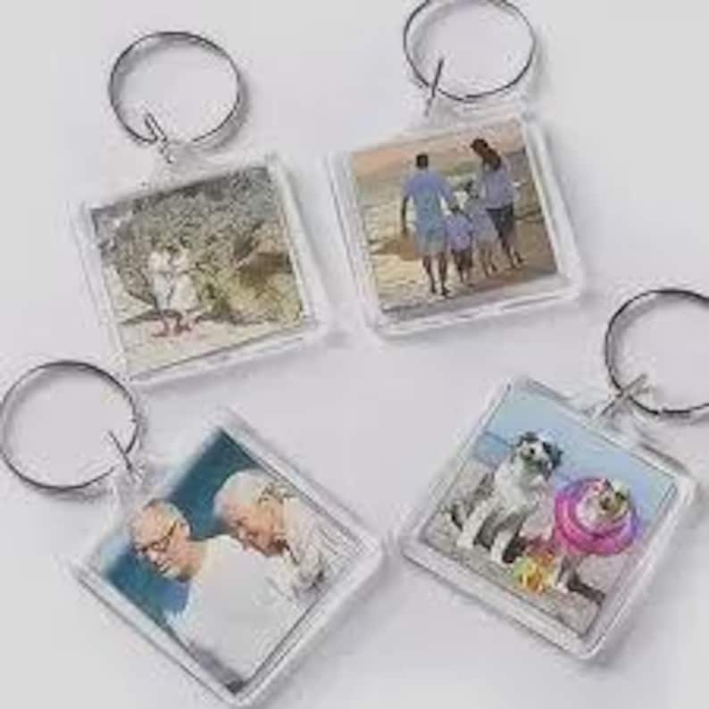 May include: Four square acrylic keychains, each featuring a different photograph insert. The images include a couple on a beach, a family walking on the sand, two older individuals, and two dogs, one wearing a pink collar, all attached to metal rings.