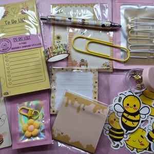 May include: A collection of bee-themed stationery and accessories. Includes a notepad, stickers, paperclips, a pen, washi tape, and a flower-shaped charm. The items are yellow, white, and black, with bee and honeycomb designs.