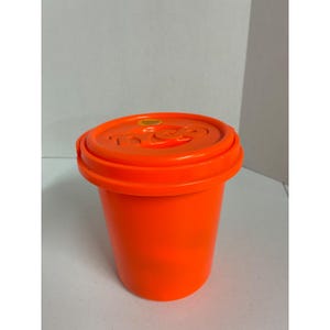 May include: A bright orange plastic storage container with a matching lid. The lid features the embossed text "tyco" in the center and a small yellow circular sticker near the edge. The container has a cylindrical shape with a wide rim.