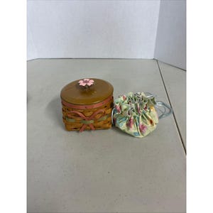 Puede incluir: Una pequeña cesta redonda tejida con tapa marrón y una flor rosa encima. La cesta está decorada con cintas rosas y verdes. Junto a ella, una pequeña bolsa floral con cordón.