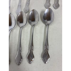 Puede incluir: Una colección de cucharas de metal plateado dispuestas en filas. Cada cuchara presenta un cuenco ovalado pulido y un mango con un patrón en relieve cerca de la base. La superficie metálica muestra un acabado reflectante y sutil.