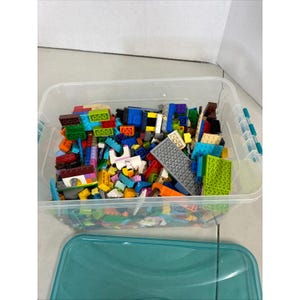 Puede incluir: Un contenedor de almacenamiento de plástico transparente lleno de una gran variedad de bloques de construcción de plástico multicolor de diversas formas y tamaños. El recipiente tiene clips de cierre color turquesa y una tapa a juego.