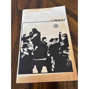 Puede incluir: Un programa vintage de "Cabaret" con una ilustración en blanco y negro de figuras. La portada presenta el título en una fuente sencilla sobre una imagen estilizada de personas en silueta. El programa está sobre una superficie de madera.