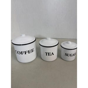Puede incluir: Tres botes de cocina de cerámica blanca con ribete y tapas negras. Los botes están etiquetados con "COFFEE", "TEA" y "SUGAR" en letras negras. Los botes son de diferentes tamaños.