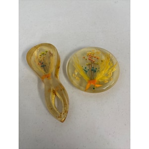 May include: Two resin pieces with dried flowers. One is a spoon-shaped object, and the other is a round, flat piece. Both are clear with yellow, orange, and blue flowers encased inside. The spoon has a hole in the handle.