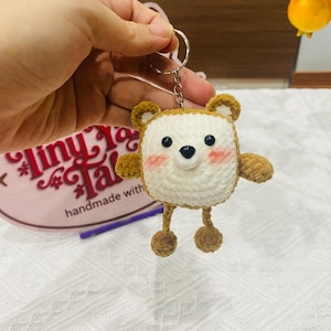 以下が含まれることがあります： 正方形の頭、白い顔、茶色のアクセントが特徴の手作りの編みぐるみクマのキーホルダー。 クマには小さな腕と脚、黒い鼻、バラ色の頬があります。 キーホルダーはシルバーの金属リングで保持されています。