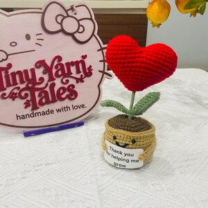Puede incluir: Una flor de corazón de ganchillo hecha a mano en una maceta. La flor roja en forma de corazón se asienta sobre un tallo verde con hojas verdes. La maceta es de color beige con una cara sonriente y una etiqueta que dice "Gracias por ayudarme a crecer."