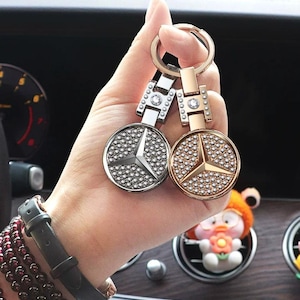 May include: Two car keyrings featuring the Mercedes-Benz logo. One is silver with a silver logo, the other gold with a gold logo. Both have a ring and are decorated with small, round, shiny embellishments, perfect for car enthusiasts.