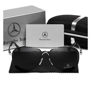 May include: Black aviator sunglasses with a silver frame, resting on a black microfiber cloth. The image includes a silver box and a black case, both with the Mercedes-Benz logo. A white card with the Mercedes-Benz logo is also visible.