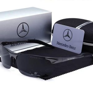 May include: Black sunglasses with the Mercedes-Benz logo on the frame and lenses. The image includes a silver box, a card, and a black case, all bearing the Mercedes-Benz emblem. The sunglasses rest on a black cloth.