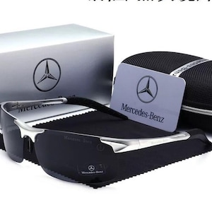 Puede incluir: Gafas de sol con montura plateada y lentes oscuras, presentadas con estuche, paño de limpieza y tarjeta. La tarjeta y la caja muestran el logotipo de Mercedes-Benz. Las gafas de sol tienen el logotipo de Mercedes-Benz en la lente.
