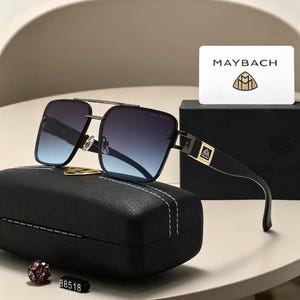 Puede incluir: Gafas de sol cuadradas con lentes degradadas azules y puente plateado. Las patillas negras presentan detalles dorados y el logotipo de Maybach. Las gafas descansan sobre un estuche negro con costuras blancas, junto a una tarjeta Maybach.