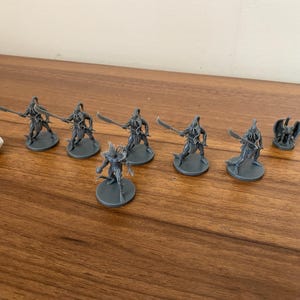 May include: A collection of gray miniature fantasy figures, including warriors with swords and a winged creature, alongside a white, multi-sided die. The figures are arranged on a wooden surface, suggesting a tabletop game setting.