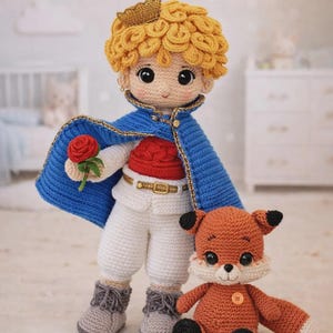 Puede incluir: Una muñeca de ganchillo de un niño con cabello amarillo rizado, corona dorada, capa azul, camisa roja, pantalones blancos y botas grises, sosteniendo una rosa roja. Junto a la muñeca hay un pequeño zorro naranja de ganchillo con detalles negros.