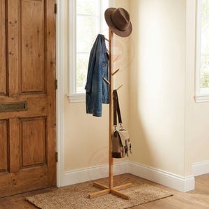 May include: A tall, light-coloured wooden coat rack with a central vertical post and a cross-shaped base. It features multiple angled pegs for hanging items, currently holding a brown fedora hat, a blue denim jacket, and a tan canvas messenger bag.