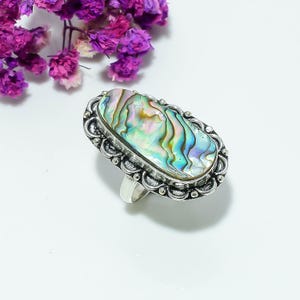 Könnte beinhalten: Ein ovaler Ring mit einer polierten, irisierenden Abalone-Muschel-Einlage in wirbelnden Grün-, Blau- und Violetttönen. Die Muschel ist in einen dekorativen, oxidierten silberfarbenen Metallrahmen mit gewelltem Rand und kleinen Perlenakzenten gefasst.