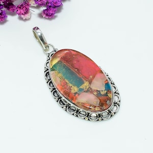 Könnte beinhalten: Ovaler Anhänger mit einem farbenfrohen Edelstein. Der Stein weist rote, rosa, goldene und blaue Farbtöne auf. Der Anhänger hat eine silberfarbene Metalleinfassung mit einem dekorativen Rand und eine Schlaufe für eine Kette.