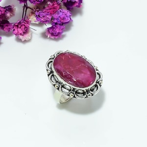 Könnte beinhalten: Ein Silberring mit einem großen, ovalen, facettierten, tiefrosa Edelstein, der in eine dekorative, gewellte Metallfassung mit poliertem Finish eingefasst ist. Die Ringschiene ist glatt und silberfarben und bildet einen Kontrast zur verzierten Fassung.