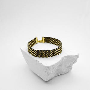 May include: A woven bead bracelet featuring a repeating checkerboard pattern of small black and metallic gold beads. The bracelet is finished with gold-toned metal end caps and a small lobster claw clasp, creating a structured band design.