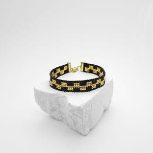 May include: A beaded bracelet featuring a repeating geometric checkerboard pattern in black and gold glass beads. The bracelet is finished with gold-toned metal end caps and a small lobster claw clasp, creating a structured and uniform band design.