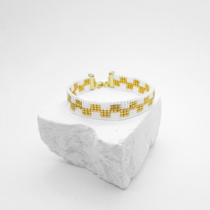 May include: A beaded bracelet featuring a repeating geometric pattern of white and metallic gold seed beads. The bracelet is finished with gold-toned metal end caps and a small lobster claw clasp, creating a structured and uniform design.