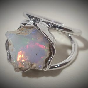 May include: A silver ring featuring a raw, uncut opal gemstone. The opal displays a play of color with flashes of red, orange, and green. The ring's band has a simple, elegant design, highlighting the natural beauty of the stone.