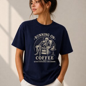 Könnte beinhalten: Ein dunkelblaues Kurzarm-T-Shirt mit einer Grafik eines Skeletts, das in einem Sessel sitzt und aus einer Tasse trinkt. Der Text lautet "RUNNING ON COFFEE AND QUESTIONABLE DECISIONS" in einem kreisförmigen Design mit Sonnenstrahlen.