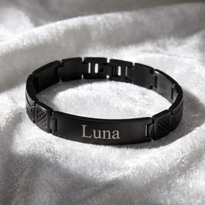 Puede incluir: Una pulsera de eslabones en negro mate con una placa rectangular central grabada con el nombre Luna en una fuente serif. La pulsera incluye detalles texturizados estilo fibra de carbono en los eslabones laterales y un cierre plegable.