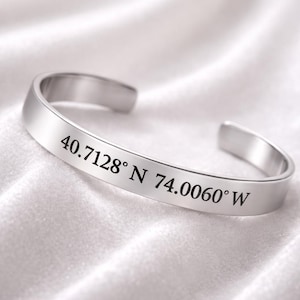 Può includere: Un bracciale a polsino argentato con le coordinate incise 40.7128° N 74.0060° W. Il bracciale è realizzato in metallo lucido e lucido ed è esposto su una superficie bianca strutturata.